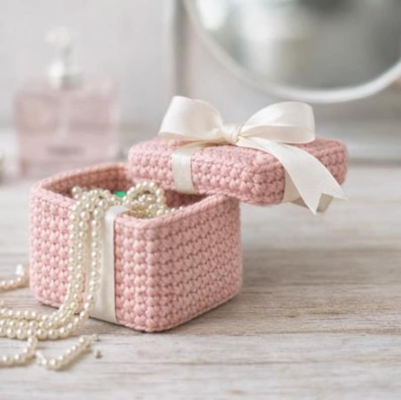 May include: A pink crocheted jewelry box with a white ribbon tied around the lid. The box is open, revealing a strand of pearls. The box is sitting on a wooden surface.