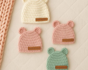 Puede incluir: Cuatro gorros de bebé hechos a mano con ganchillo en crema, rosa y verde, cada uno con orejas de oso. Los gorros tienen un punto texturizado y una pequeña etiqueta rectangular que dice "HearthCraft Studio Co.".