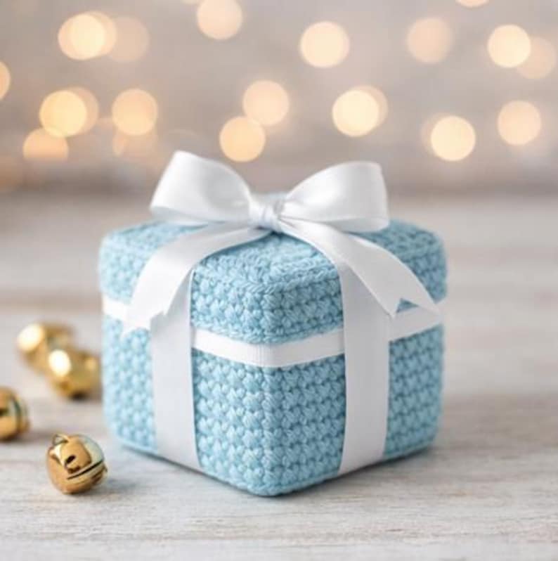 May include: A light blue, crocheted gift box tied with a white satin ribbon. The square box has a textured appearance and is decorated with a large bow on top. The background features soft, blurred lights.