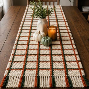 Puede incluir: Un camino de mesa rectangular de ganchillo con una base de color crema y un patrón de cuadrícula formado por líneas entrecruzadas de color verde oscuro y naranja quemado. El camino termina con largos flecos alternos en crema, verde y naranja.