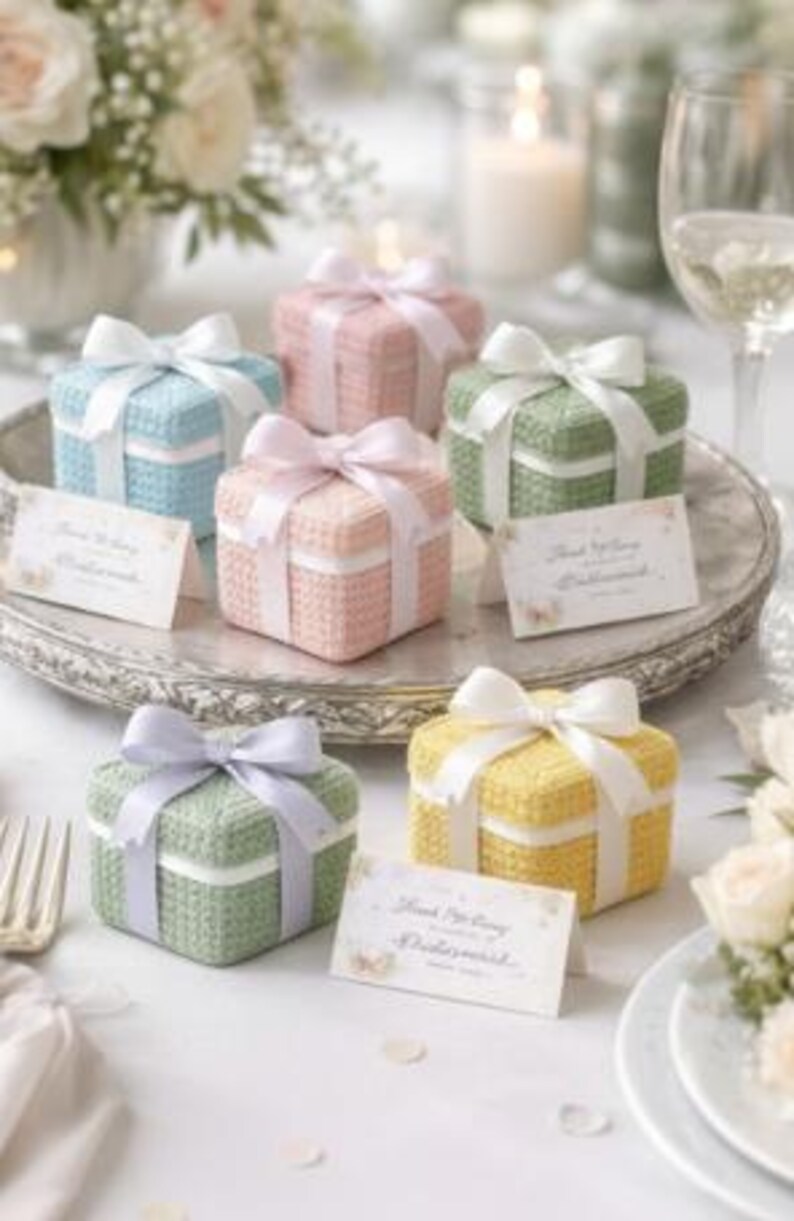 May include: A collection of small, square gift boxes in pastel colors: blue, pink, green, and yellow. Each box is tied with a matching satin ribbon bow. The boxes are arranged on a silver tray, with place cards and floral decorations.
