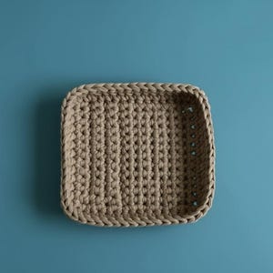 May include: A beige, square crochet basket with rounded corners. The basket is made of thick, textured yarn and has a solid, woven base. The basket is empty and sits on a solid blue background.