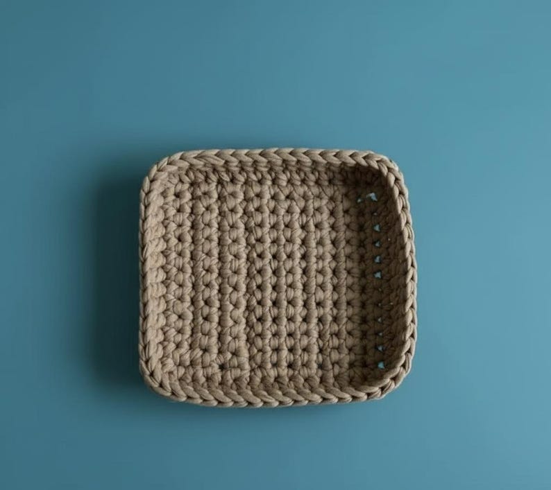 May include: A beige, square crochet basket with rounded corners. The basket is made of thick, textured yarn and has a solid, woven base. The basket is empty and sits on a solid blue background.