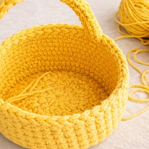 Op de afbeelding: Een handgemaakte gele gehaakte mand met een handvat. De ronde mand heeft een getextureerd oppervlak. Twee bollen geel garen en los garen liggen naast de mand. Perfect voor opslag of decoratie.