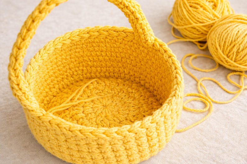 Op de afbeelding: Een handgemaakte gele gehaakte mand met een handvat. De ronde mand heeft een getextureerd oppervlak. Twee bollen geel garen en los garen liggen naast de mand. Perfect voor opslag of decoratie.