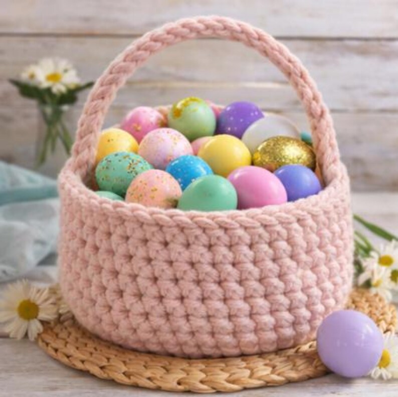 Op de afbeelding: Een roze gehaakte paasmand gevuld met kleurrijke gespikkelde paaseieren. De eieren zijn in verschillende kleuren, waaronder roze, blauw, geel, groen en paars. De mand heeft een gevlochten handvat en staat op een geweven mat.