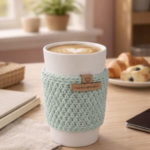 May include: A white ceramic mug with a light green crocheted cozy, holding a latte with a heart-shaped design. The cozy has a small leather tag that reads "HearthCraftStudio". The mug is on a wooden table.