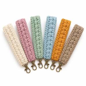 May include: Six wristlet keychains arranged in a fan shape, each featuring a textured macrame cord design in cream, dusty rose, sage green, light blue, mustard yellow, and tan. Each keychain includes a bronze-toned metal lobster clasp.