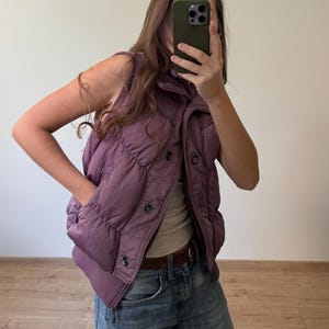 May include: A mauve sleeveless puffer vest featuring a high collar, a front button closure, and a ribbed hem. The vest is worn over a neutral-colored top and blue denim jeans with a brown leather belt.