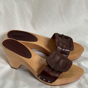 Puede incluir: Un par de sandalias de cuero marrón oscuro con tacón de cuña y suela de plataforma de madera. Cada sandalia presenta una tira ancha de cuero en la parte superior con una hebilla grande a juego y pequeños remaches plateados en la base.