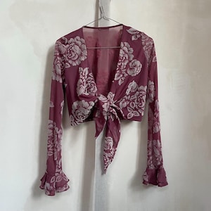 May include: A burgundy sheer top with long sleeves and a tie front closure. The top features a floral pattern in a lighter shade of burgundy. The sleeves have a flared ruffle at the wrist. The top is hanging on a metal hanger.