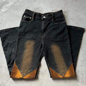 May include: Dark wash denim jeans with a high waist and flared legs. The jeans feature a faded brown design on the front and brown suede-like patches at the bottom of the legs. The jeans have a classic five-pocket design.