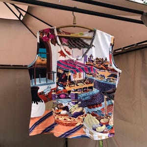 Puede incluir: Una camiseta sin mangas blanca con un estampado vibrante y colorido que representa una escena de mercado. La obra de arte incluye puestos, cestas de productos y detalles arquitectónicos. La camiseta está colgada de una percha de madera.