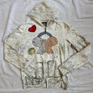 May include: A cream-colored zip-up hoodie featuring a hood with a sheer lace overlay. The front displays a central graphic of a stylized creature, a red sequined heart, and floral motifs. The fabric is covered in a fine, dark line-art leaf pattern.
