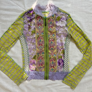 Puede incluir: Una chaqueta de manga larga con cremallera que combina paneles de encaje floral en verde lima y lavanda, mangas a rayas y detalles laterales de lunares negros. La parte frontal incluye una cremallera central verde lima y lentejuelas.
