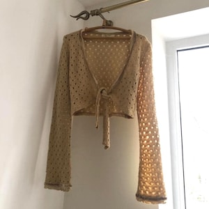 May include: A tan, open-front, long-sleeved crochet cardigan with a tie closure. The cardigan is hanging on a wooden hanger. The sleeves have a flared design and are trimmed with a darker brown color.