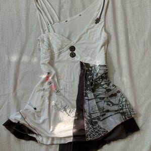May include: A white sleeveless top featuring an asymmetrical neckline with multiple thin straps, two dark circular buttons, and a side panel with a black and gray abstract splatter print. The garment includes a layered hem with sheer black fabric.