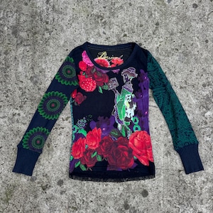 May include: A long-sleeve shirt with a black base featuring a vibrant graphic print of red roses and a figure in a green kimono holding a book. The left sleeve has green circular patterns, the right sleeve has green leafy designs, and the hem has black lace.