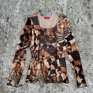 May include: A long-sleeve mesh top featuring a brown, black, and beige geometric pattern. The garment has a scoop neckline with a beige inner lining and a red label reading TAIFUN. A central floral-shaped appliqué is embellished with silver sequins.