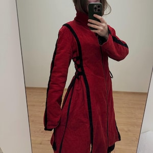 May include: A long, red coat with black trim and a high collar. The coat has black accents on the sleeves and a corset-style back. The coat is made of a textured fabric.
