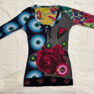 Pode incluir: Uma blusa de manga comprida com um design único. A blusa apresenta uma mistura de padrões, incluindo círculos azuis, estampas florais e uma grande rosa vermelha. O decote tem um cordão e a bainha inferior é preta com detalhes florais.