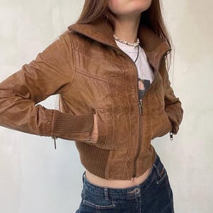 May include: A brown leather jacket with a zip-up front, ribbed cuffs, and a ribbed hem. The jacket has a high collar and two front pockets. The jacket is worn over a white t-shirt and blue jeans.