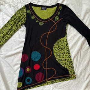 May include: Black long-sleeve tunic with a V-neck and colorful accents. The tunic features a green patterned sleeve and a green panel with a bubble design. It has a series of colorful circles and orange stitching details.