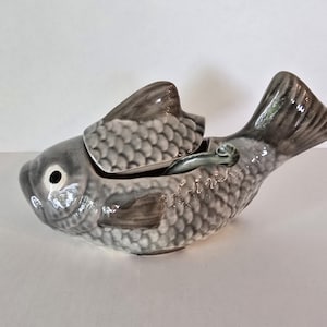 May include: A ceramic fish-shaped lidded container with a glossy grey and white glaze. The body features detailed scale textures, a prominent dorsal fin lid, and a curved tail. A small matching ceramic spoon rests inside the open top section.