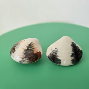 May include: Two ceramic clam-shaped objects with a glossy off-white finish. Each piece features hand-painted, abstract brushstrokes in shades of dark brown, charcoal gray, and muted copper, creating a distinct pattern on the curved surfaces.