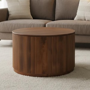 Op de afbeelding: Een ronde, bruine salontafel met verticale groeven. De tafel staat op een wit tapijt voor een bruine bank met twee decoratieve kussens. Een potplant en een vloerlamp staan ook in de kamer.