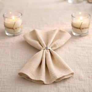 May include: A folded beige linen napkin with a silver ring, on a beige tablecloth. Two lit candles in glass holders with twine accents are either side of the napkin, creating a warm table setting.