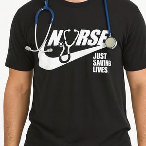 Op de afbeelding: Zwart t-shirt met een witte opdruk van "Nurse" met een stethoscoopontwerp en de woorden "Just Saving Lives". Een blauwe stethoscoop wordt om de nek gedragen.