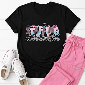 May include: Black t-shirt with the words "STAFFING coordinator" in a colorful design, paired with pink scrub pants and white canvas sneakers. The shirt's design includes medical symbols and a leopard print pattern.
