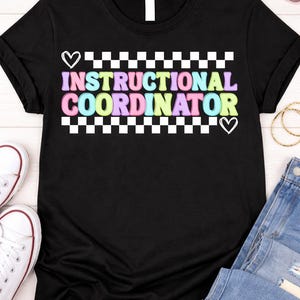 May include: Black t-shirt with the words "INSTRUCTIONAL COORDINATOR" in colorful, bubble letters. The text is framed by a white and black checkered pattern and small heart shapes. The shirt is displayed with jeans and sneakers.