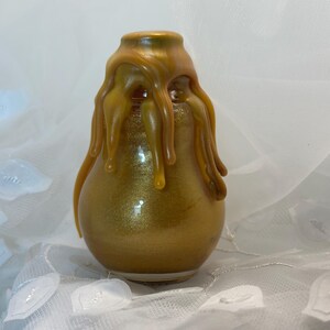 May include: A pear-shaped, gold-colored vase with a metallic finish. The top of the vase is adorned with a dripping, amber-colored design. The vase sits on a white, textured surface.