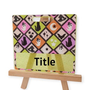 May include: A square sign with a diamond pattern featuring ghosts, black cats, bats, moons, and stars in pink, green, and black. The sign has a gold glitter section with the word Title printed in black, resting on a small wooden easel stand.