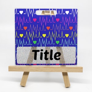 May include: A blue and white name tag with a silver glitter background. The tag has a pattern of colorful hearts and EKG lines. The text "Title" is printed in black on the tag. The tag is displayed on a small wooden easel.