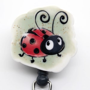 May include: A white, round badge holder with a cartoon ladybug design. The ladybug is red with black spots and has black eyes. The badge holder has a gray retractable cord.