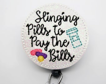 May include: White round badge with glitter and black text that reads "Slinging Pills to Pay the Bills". There is a small blue and white pill bottle and a few colorful pills on the badge.