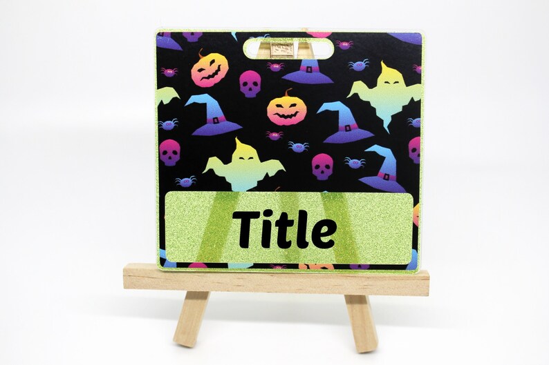 K&ouml;nnte beinhalten: Ein Halloween-Themen-Ausweishalter mit schwarzem Hintergrund, der bunte K&uuml;rbisse, Hexenh&uuml;te, Geister, Totenk&ouml;pfe und Spinnen zeigt. Der Ausweishalter hat einen gr&uuml;n glitzernden Bereich mit dem Wort "Title" in Schwarz. Er wird auf einer kleinen Holzstaffelei pr&auml;sentiert.