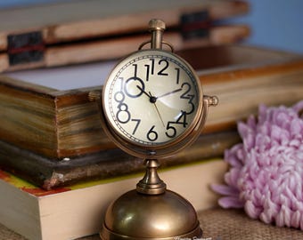May include: A brass-colored desk clock with a cream-colored face and black numerals. The clock has a round face and a decorative base. The hands point to approximately 10:10. The clock is set on a table next to books and a pen.