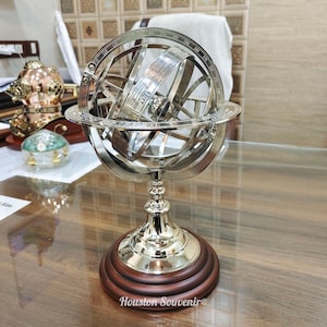 May include: A silver armillary sphere with a zodiac band, set on a wooden base. The sphere features multiple rings and a central globe. The base is dark brown with a Houston Souvenir logo.