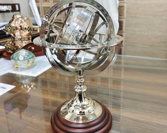 May include: A silver-colored armillary sphere with a wooden base. The sphere has multiple rings and a central globe, with the word "ARIES" visible. The base is dark brown, and the text "Houston Souvenir" is printed on the base.