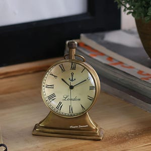 Peut inclure: Une horloge ronde de couleur laiton avec des chiffres romains et le mot "London" sur le cadran. Un symbole d'ancre est au-dessus du mot. L'horloge repose sur une base rectangulaire. L'horloge est sur une surface en bois.