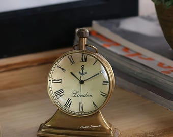 May include: A brass-colored, round clock with Roman numerals and the word "London" on the face. An anchor symbol is above the word. The clock sits on a rectangular base. The clock is on a wooden surface.