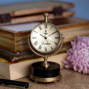 Könnte beinhalten: Eine messingfarbene Tischuhr mit cremefarbenem Zifferblatt und schwarzen römischen Ziffern. Das Zifferblatt zeigt ein Ankersymbol und das Wort "London". Die Uhr steht auf einem schwarzen Sockel mit goldenen Akzenten. Durchmesser ca. 10 cm.