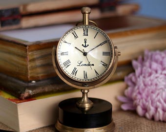 May include: A brass-colored desk clock with a cream-colored face and black Roman numerals. The clock face features an anchor symbol and the word "London". The clock sits on a black base with gold accents.
