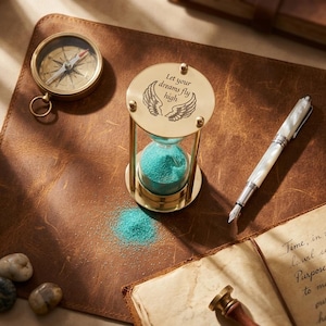 Könnte beinhalten: Eine Messing-Sanduhr mit türkisfarbenem Sand, auf deren Oberseite "Let your dreams fly high" und Flügelillustrationen eingraviert sind. Ein Kompass, ein Stift und ein offenes Buch befinden sich ebenfalls auf der braunen Lederoberfläche.