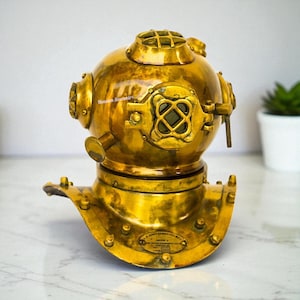 Puede incluir: Un casco de buceo de latón vintage con una parte superior redonda y una base acampanada. El casco presenta múltiples portillos, remaches y una ventana de visualización frontal. El latón tiene un acabado dorado pulido, y el casco es un artículo náutico decorativo.