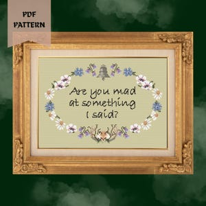 May include: A cross stitch pattern in a gold frame featuring the text Are you mad at something I said? surrounded by an oval wreath of white and blue flowers, a small grey bell at the top, and a fox head with antlers at the bottom center.